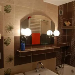 Wykończona łazienka z brązowymi i beżowymi płytkami, wzorami liści na ścianach, dużym lustrem z czterema okrągłymi lampami, umywalką i wanną.