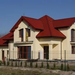 Gotowy, dwukondygnacyjny dom jednorodzinny z czerwonym dachem, kremową elewacją i ogrodzeniem z siatki.