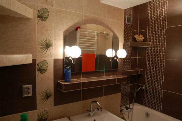 Wykończona łazienka z brązowymi i beżowymi płytkami, wzorami liści na ścianach, dużym lustrem z czterema okrągłymi lampami, umywalką i wanną.