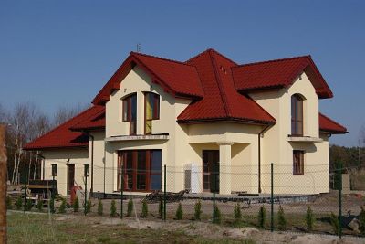 Gotowy, dwukondygnacyjny dom jednorodzinny z czerwonym dachem, kremową elewacją i ogrodzeniem z siatki.
