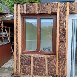 PIOTR TOKARCZYK - Montaż okna z izolacją z wełny mineralnej, widoczne drewniane elementy konstrukcyjne i odbicie osoby robiącej zdjęcie w szybie okna.