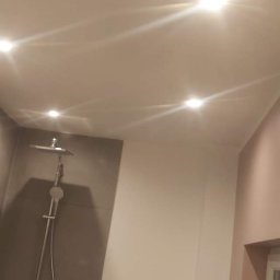 PIOTR TOKARCZYK - Wykończona łazienka z szarą glazurą, białą armaturą i oświetleniem punktowym. Widoczny prysznic, toaleta i okno. Ściany w odcieniach szarości i delikatnego różu.