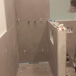 PIOTR TOKARCZYK - Wnętrze kabiny prysznicowej w trakcie wykańczania, z szarymi płytkami i widocznymi elementami instalacji wodnej oraz narzędziami pracy na podłodze.