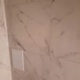 Wykończona ściana z płytek imitujących marmur w odcieniach bieli i szarości. Widoczny rewizyjny właz. Minimalistyczne wykończenie łazienki.