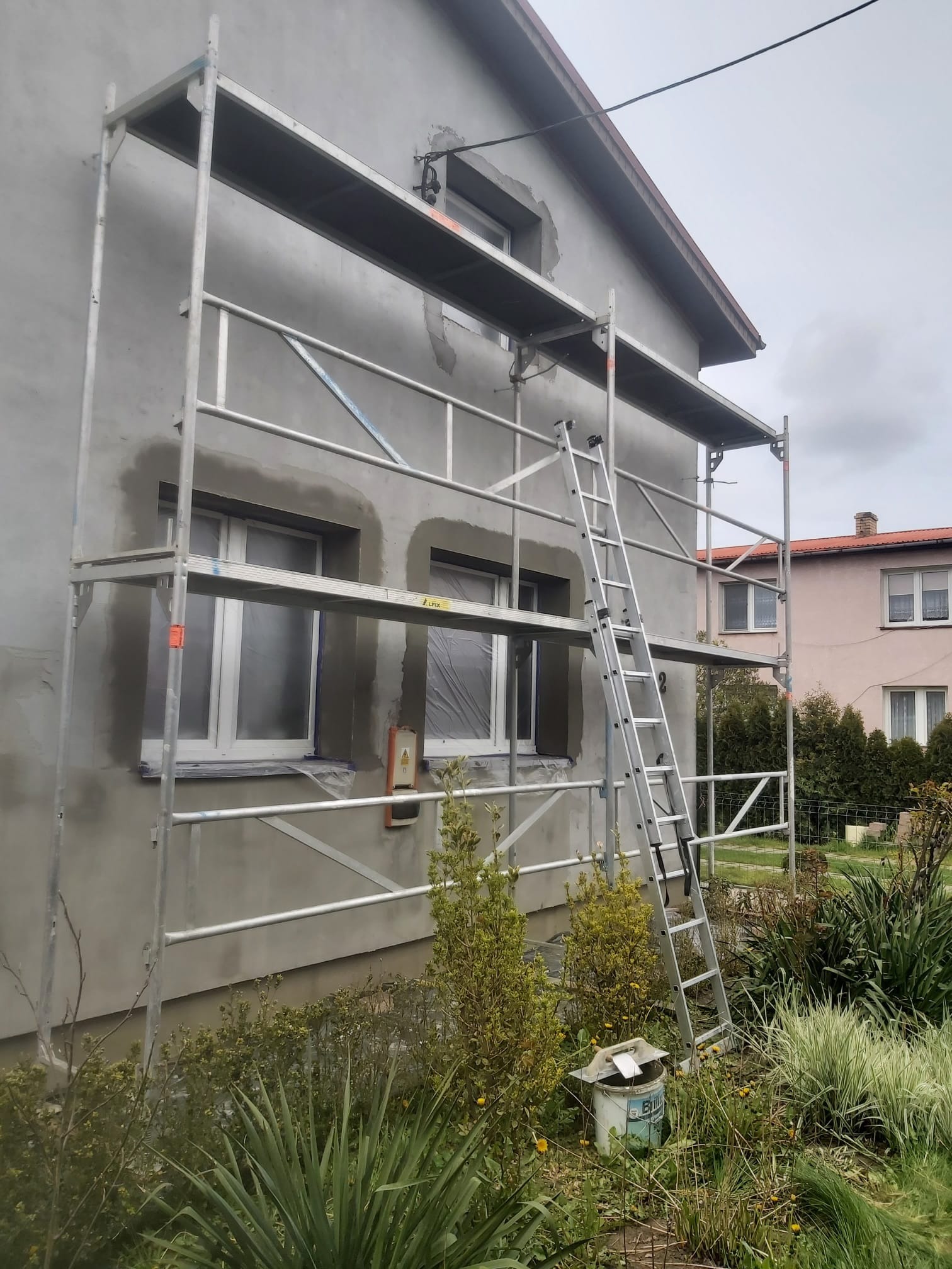 Dom w trakcie szpachlowania elewacji. Widoczne rusztowanie, okna zabezpieczone folią, ślady po szpachlowaniu. Na pierwszym planie roślinność.