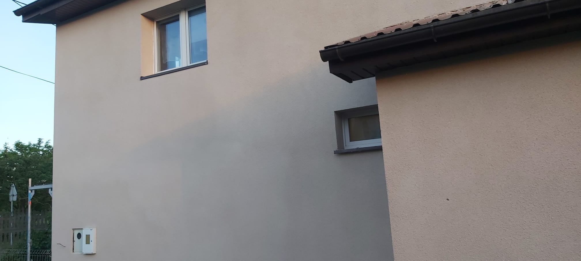 Fragment elewacji domu po remoncie: gładki tynk w odcieniach beżu, okna z białymi ramami i ciemny dach z rynną. Widoczna skrzynka elektryczna na ścianie.