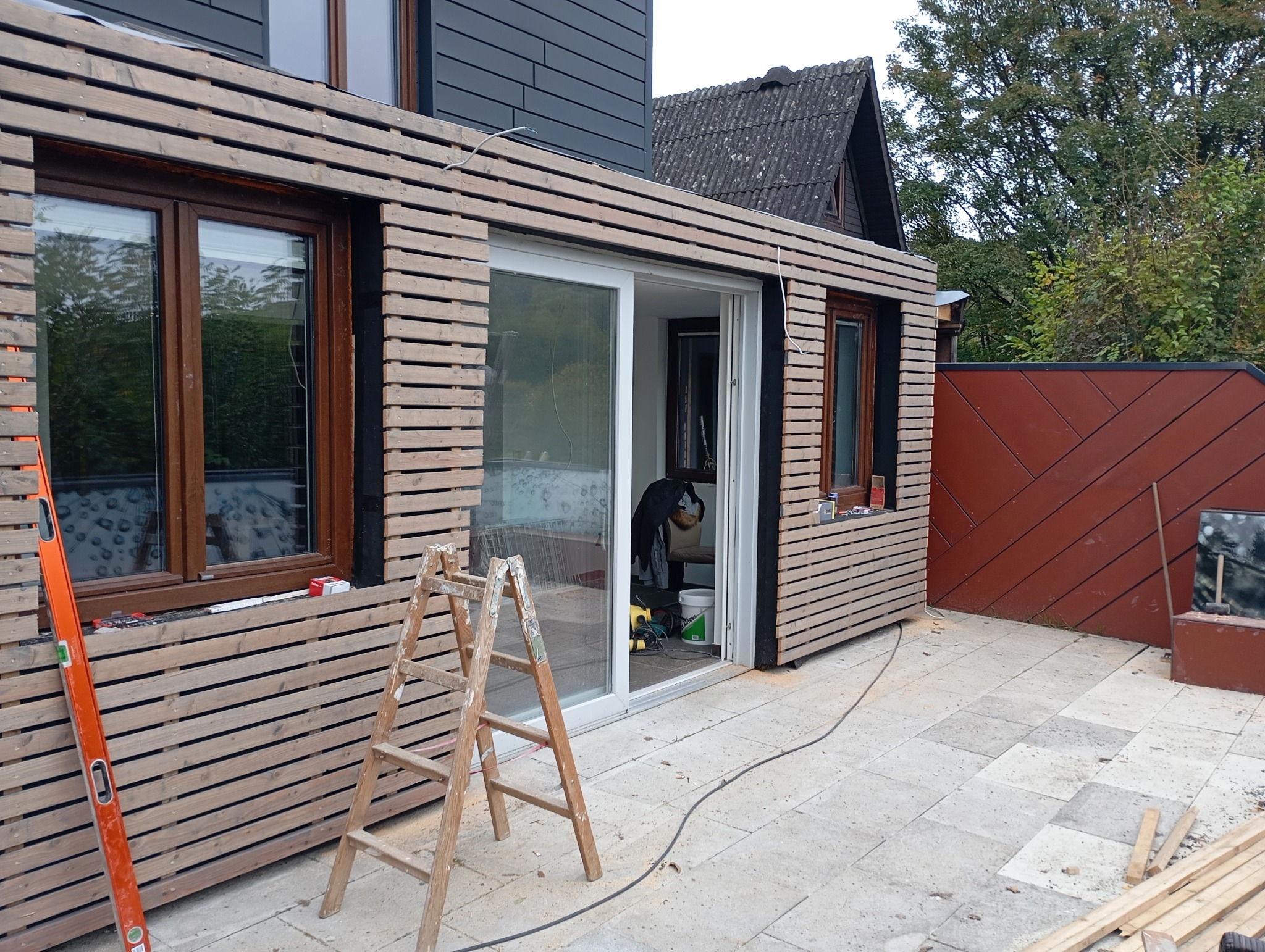 Montaż drewnianej elewacji na budynku z oknami i przesuwnymi drzwiami. Widoczna drabina, poziomica i narzędzia. Etap prac wykończeniowych.