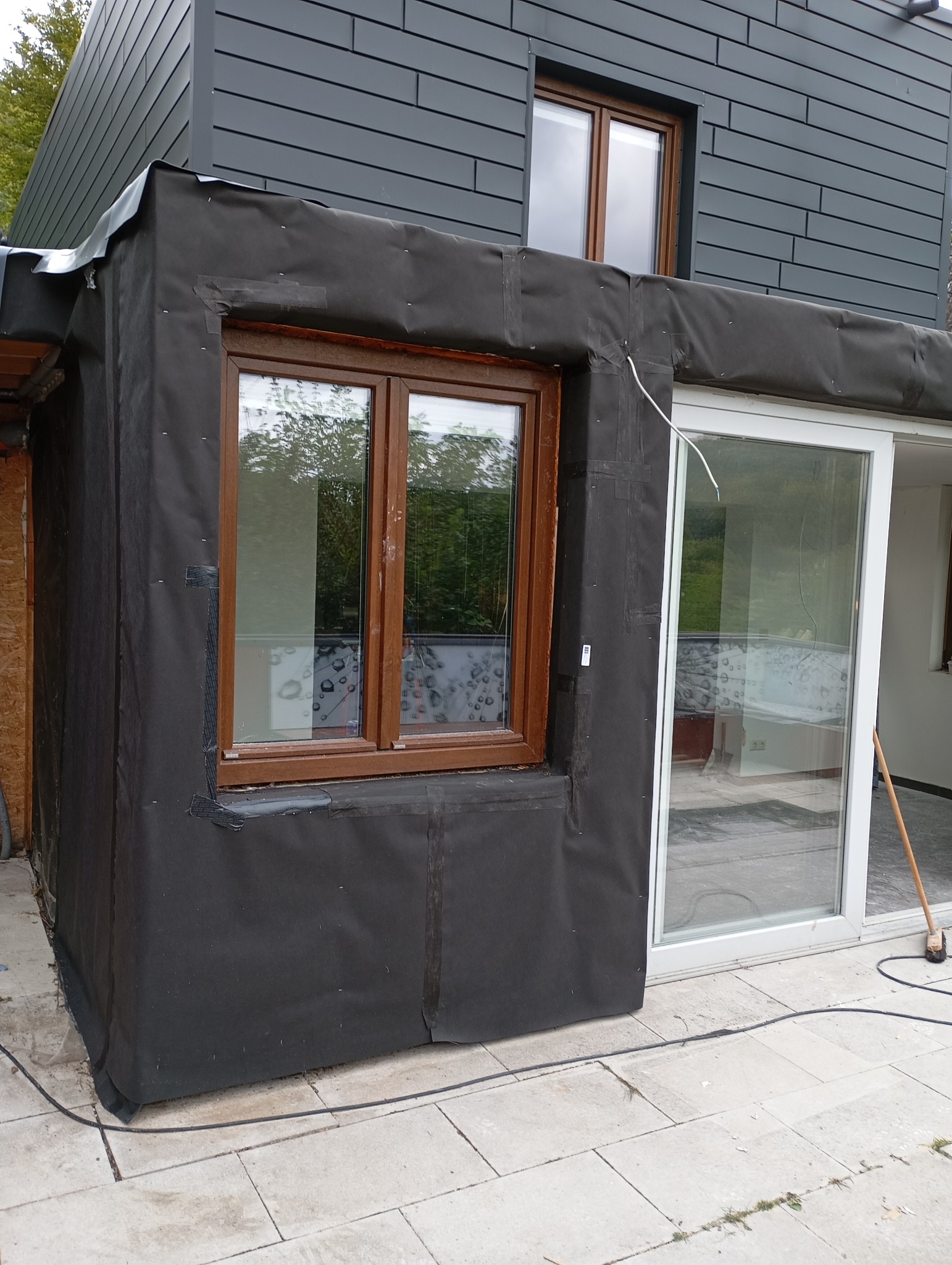 Etap prac wykończeniowych elewacji domu: okno z brązową ramą zabezpieczone czarną folią budowlaną, widoczna część elewacji z szarymi panelami, okno przesuwne.