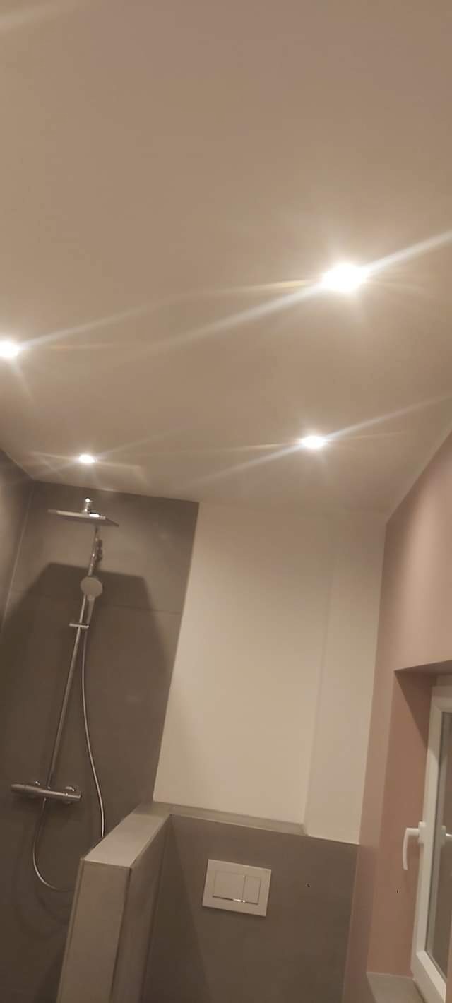 Wykończona łazienka z szarą glazurą, białą armaturą i oświetleniem punktowym. Widoczny prysznic, toaleta i okno. Ściany w odcieniach szarości i delikatnego różu.