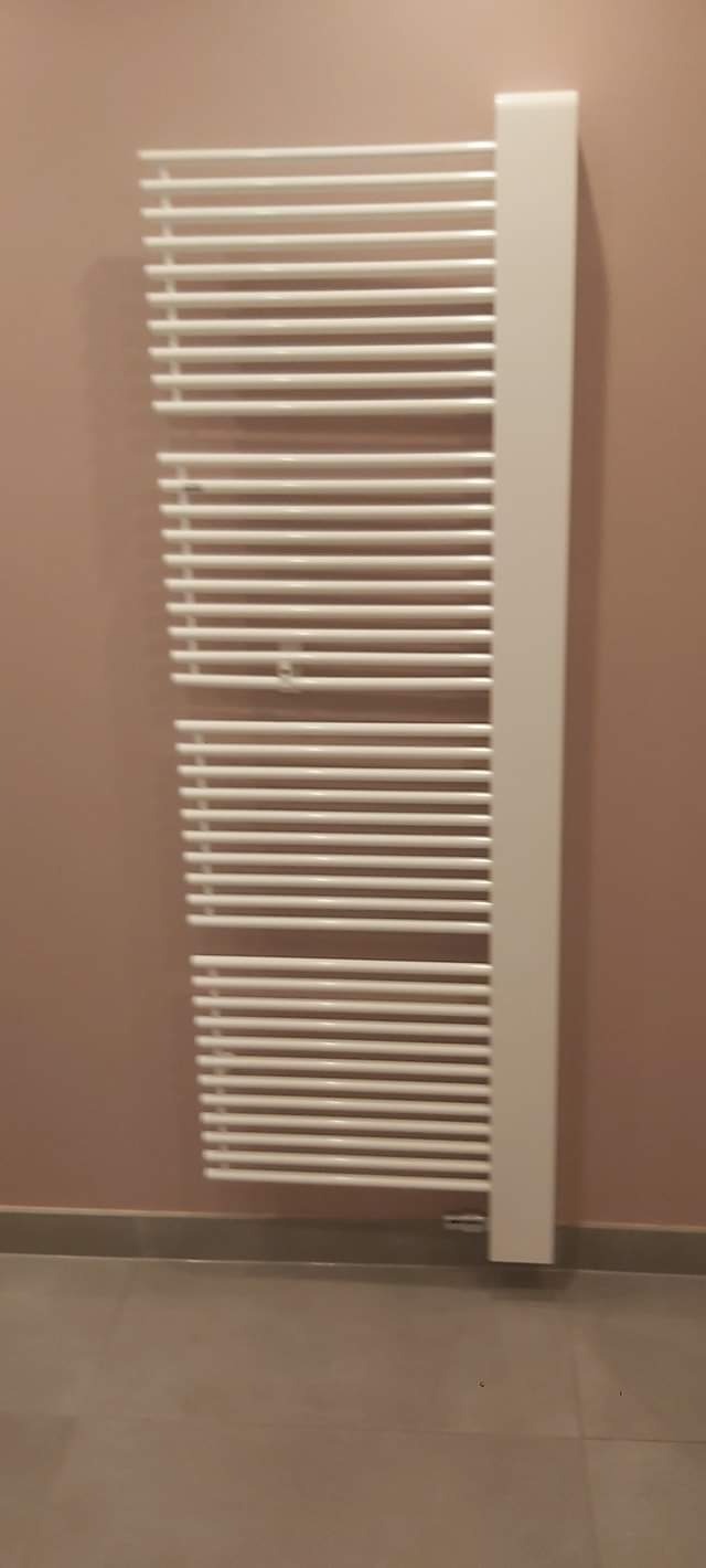 Biały, drabinkowy grzejnik łazienkowy zamontowany na pudrowo-różowej ścianie, widoczny fragment szarej podłogi. Nowoczesny design, ciepłe wnętrze.