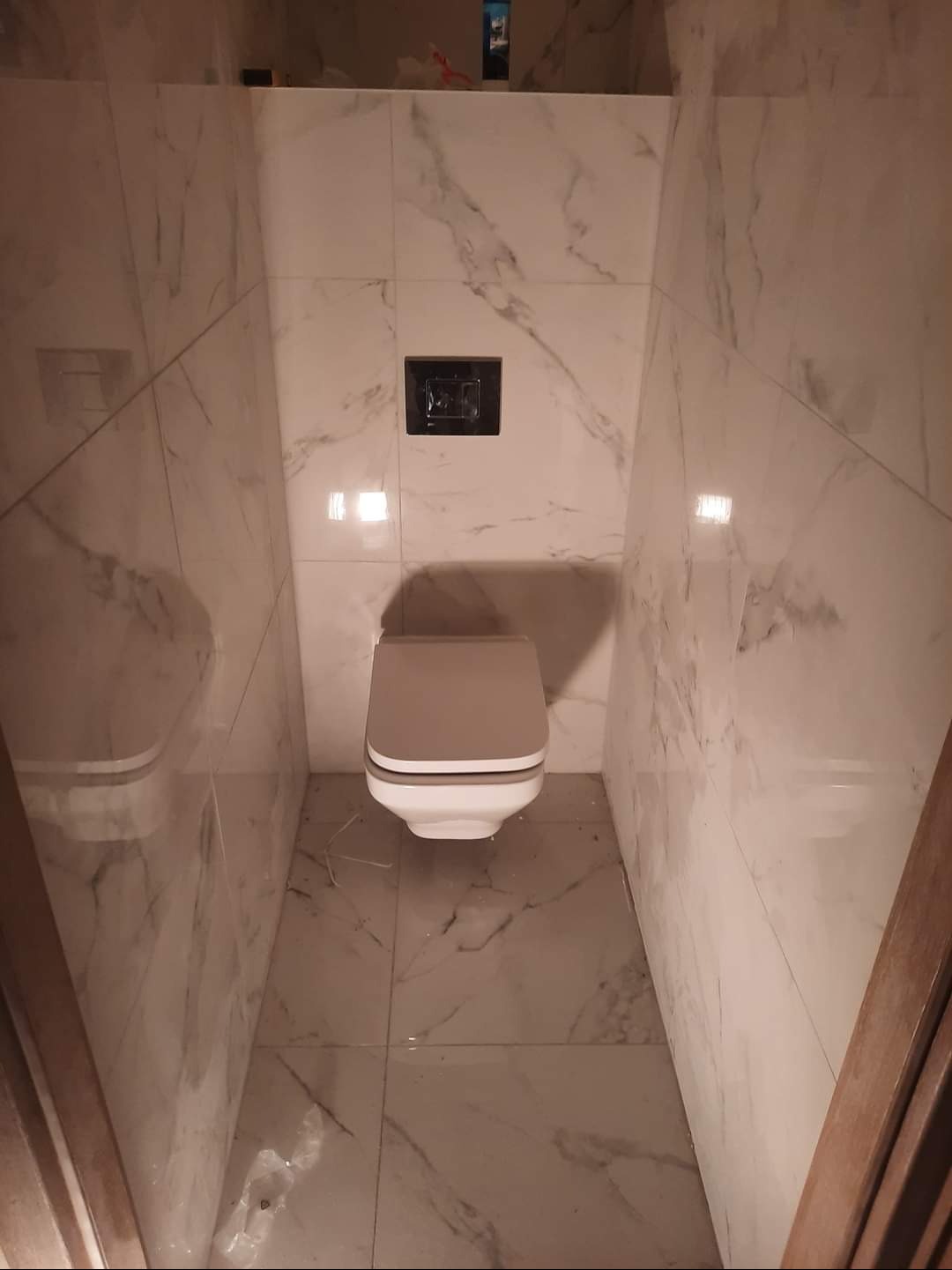 Kompakt WC z szarą deską i czarnym przyciskiem spłukującym, otoczony marmurowymi płytkami podłogowymi i ściennymi. Widok z góry, wąska przestrzeń.