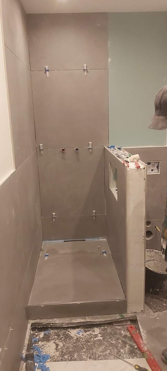 Wnętrze kabiny prysznicowej w trakcie wykańczania, z szarymi płytkami i widocznymi elementami instalacji wodnej oraz narzędziami pracy na podłodze.