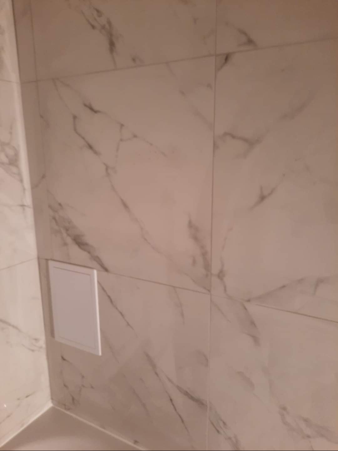 Wykończona ściana z płytek imitujących marmur w odcieniach bieli i szarości. Widoczny rewizyjny właz. Minimalistyczne wykończenie łazienki.