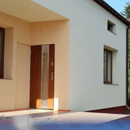 Ocieplony dom jednorodzinny z brązowymi oknami i drzwiami wejściowymi. Widoczna elewacja w kolorze kremowym i fragment brukowanego tarasu. Prace wykończeniowe.