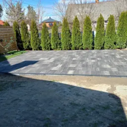 Ułożona kostka brukowa w geometryczny wzór, obramowana czarnym brzegiem. W tle rząd tuj i drewniana balia, sugerujące strefę relaksu w ogrodzie.