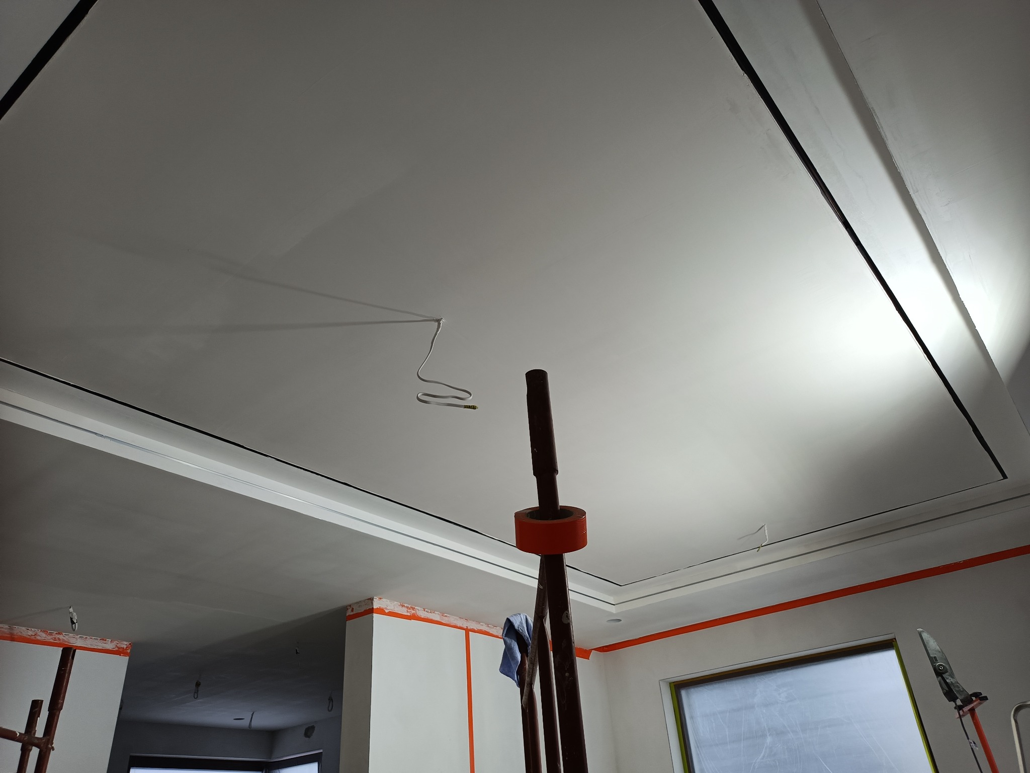 Biały sufit z dekoracyjnymi listwami i wystającym kablem, zabezpieczone ściany taśmą malarską, fragment rusztowania w pomieszczeniu w trakcie wykończenia.