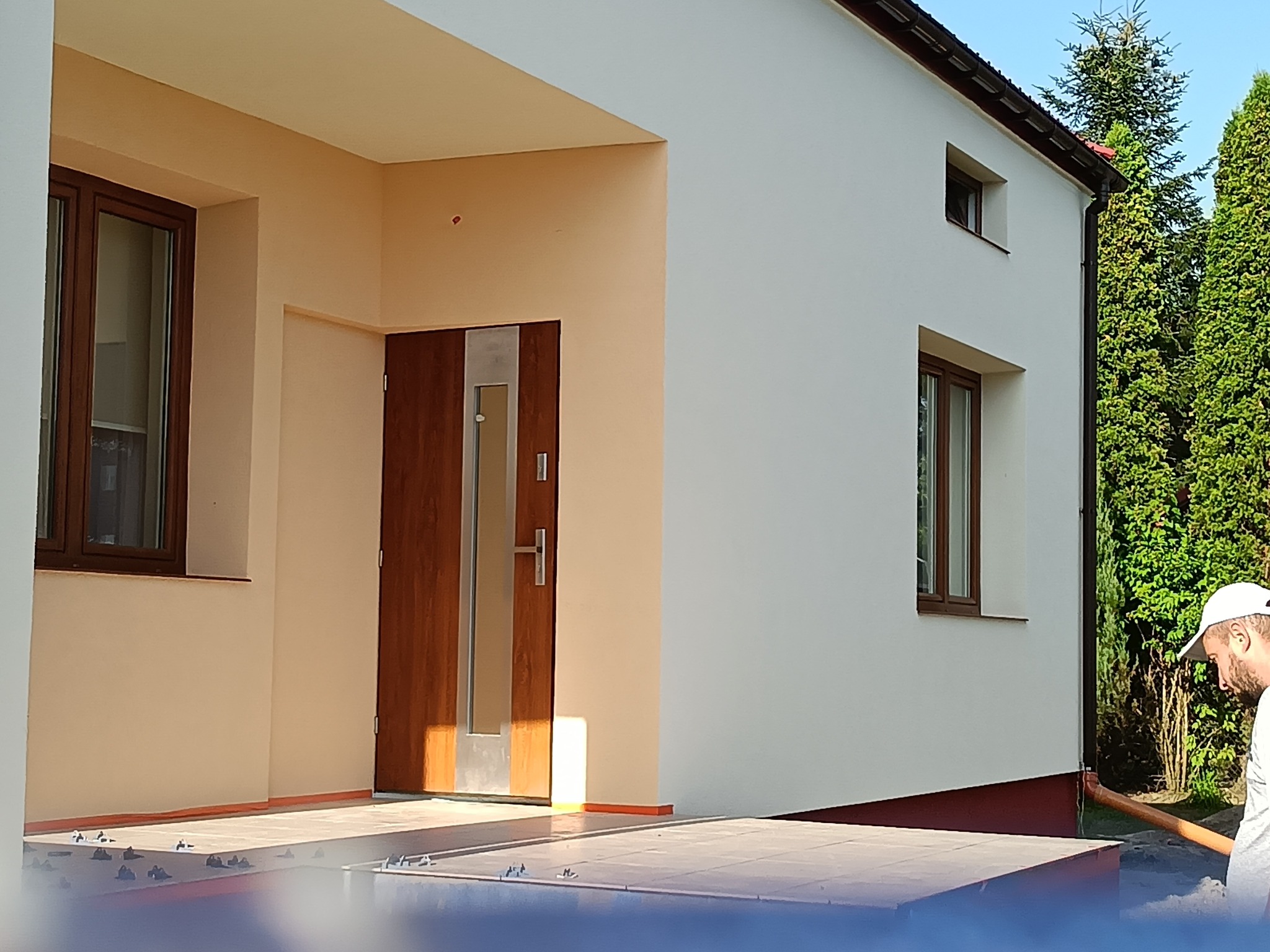 Ocieplony dom jednorodzinny z brązowymi oknami i drzwiami wejściowymi. Widoczna elewacja w kolorze kremowym i fragment brukowanego tarasu. Prace wykończeniowe.