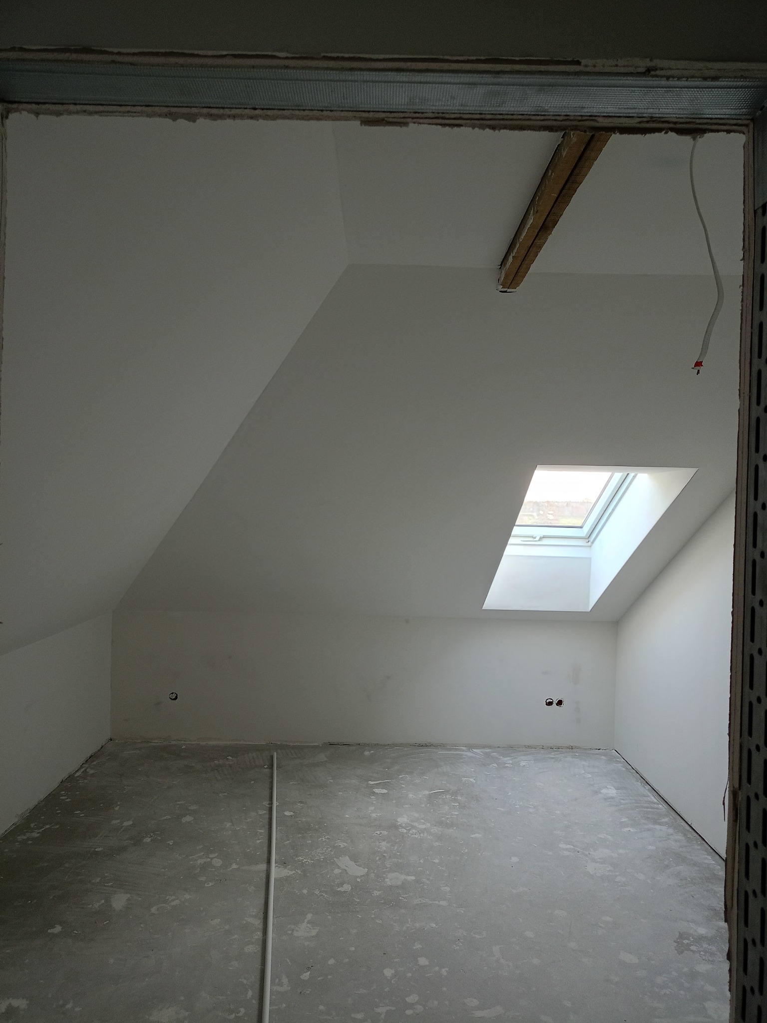 Poddasze po remoncie z oknem dachowym, świeżo pomalowane ściany na biało, surowa wylewka. Widoczne elementy konstrukcyjne sufitu i ściany. Wnętrze w stanie surowym.