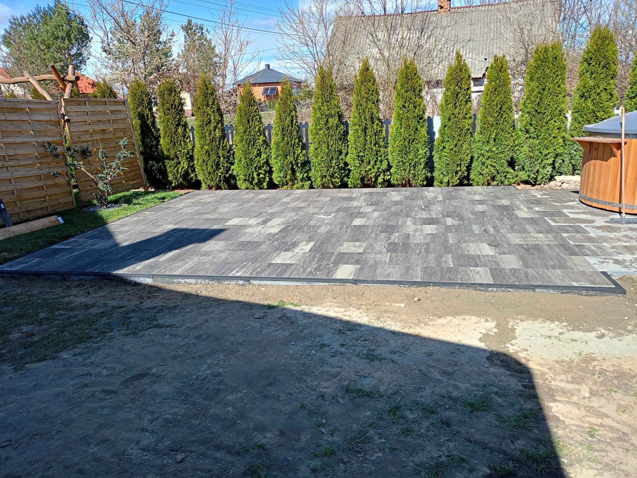 Ułożona kostka brukowa w geometryczny wzór, obramowana czarnym brzegiem. W tle rząd tuj i drewniana balia, sugerujące strefę relaksu w ogrodzie.