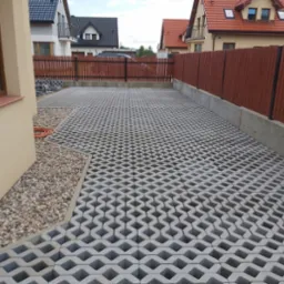 Ułożona szara ażurowa kostka brukowa tworząca podjazd na posesji, widok z boku budynku, z brązowym ogrodzeniem w tle i kamieniami ozdobnymi po bokach.