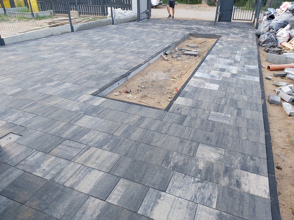Układanie kostki brukowej w szaro-czarnych odcieniach, z widocznym miejscem na rabatę. Część kostki jeszcze nieułożona, widoczne materiały budowlane.