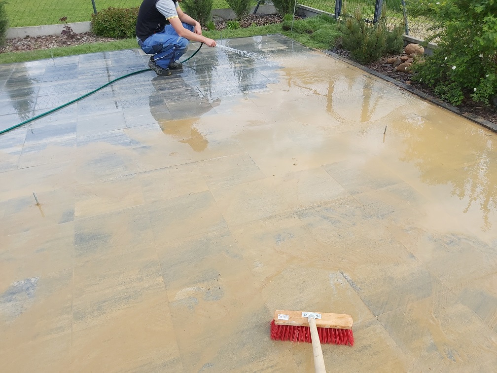 Czyszczenie kostki brukowej po ułożeniu. Mężczyzna spłukuje wodą nową nawierzchnię z szarej kostki brukowej. Na dole kadru leży czerwona szczotka.