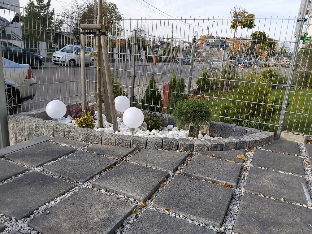 Ścieżka z kwadratowych płyt brukowych, wysypana drobnym białym żwirem, obok rabata z kamienia z ozdobnymi krzewami i trzema białymi kulami-lampami.
