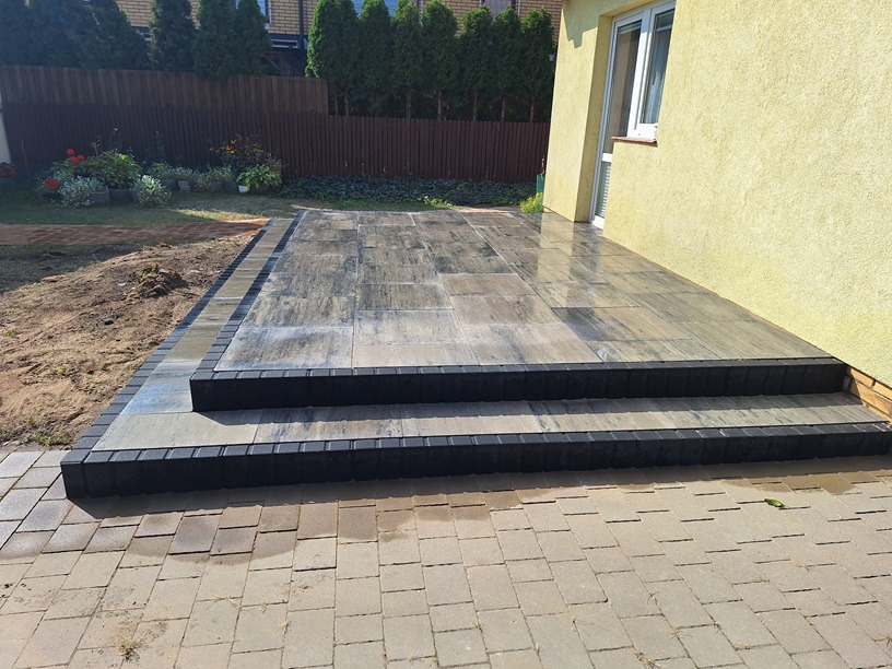 Wykończony taras z kostki brukowej w odcieniach szarości, z dwoma stopniami obrzeżonymi ciemną kostką, przylegający do budynku z oknem i zielonym tłem z tujami.