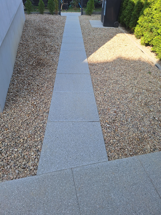 Ścieżka z szarych płyt betonowych, otoczona ozdobnym żwirem, prowadząca wzdłuż budynku.  Widoczne tuje i kosz na śmieci w tle.