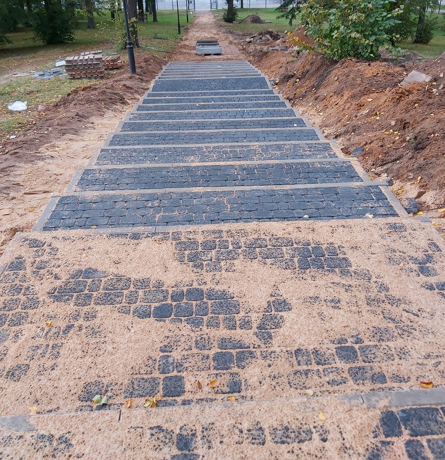 Schody z kostki brukowej w trakcie budowy, widok z dołu. Częściowo zasypane piaskiem, w tle drzewa i latarnie. Widoczny etap prac brukarskich.