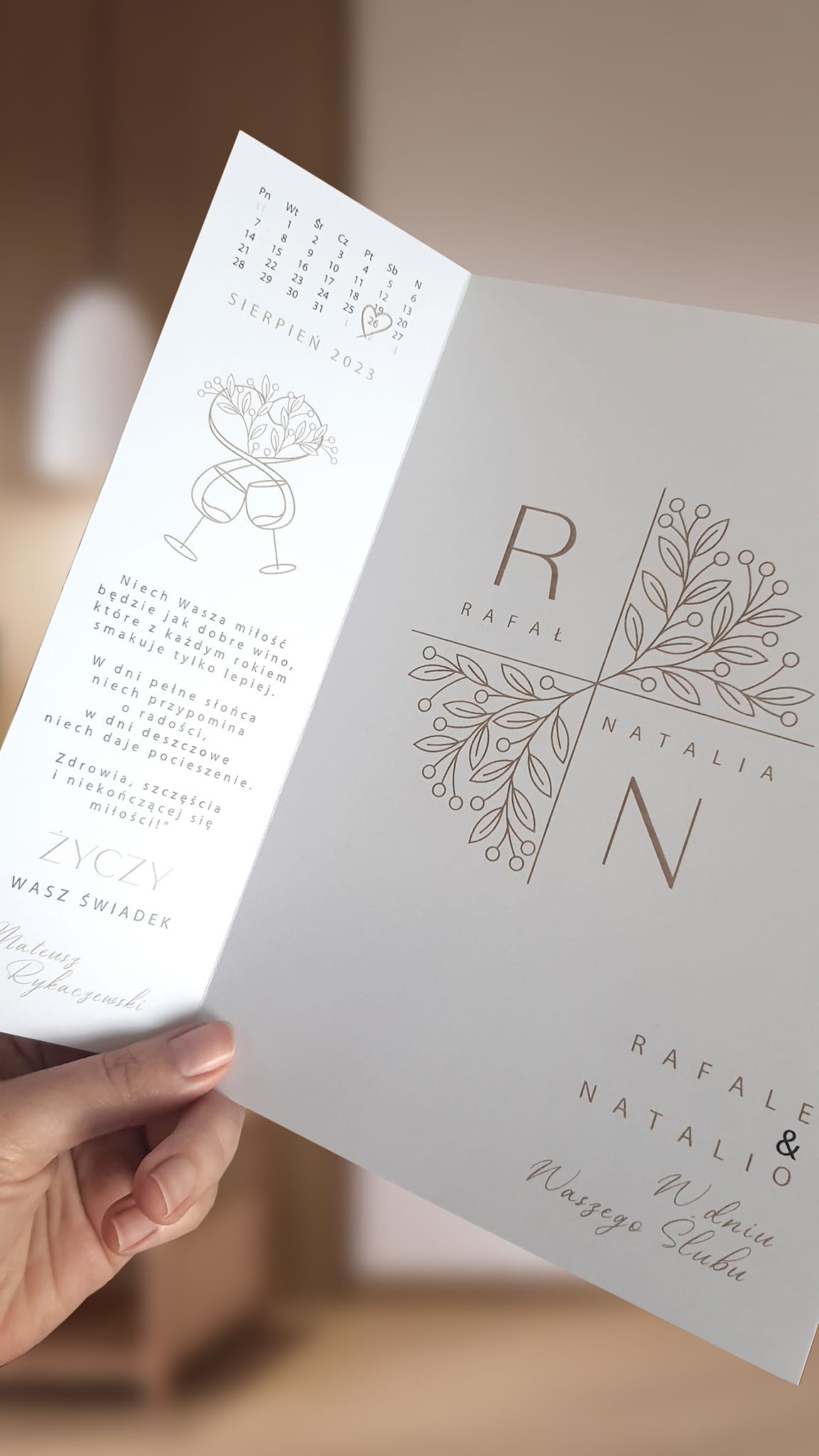Otwarta, kremowa kartka okolicznościowa z kalendarzem sierpnia 2023 i życzeniami ślubnymi, trzymana w dłoni, z widocznymi imionami Rafał i Natalia.