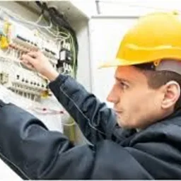 Elektryk w kasku łączy przewody w skrzynce rozdzielczej. Widoczne bezpieczniki i automatyka. Praca precyzyjna, w tle ściana, oświetlenie jasne.