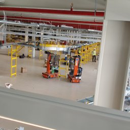 energon - Wnętrze nowoczesnej hali z rozbudowanymi instalacjami: czerwone rury przeciwpożarowe, korytka kablowe, system przenośników. Widoczne mobilne podnośniki i pracownik.