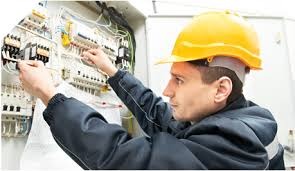 Elektryk w kasku łączy przewody w skrzynce rozdzielczej. Widoczne bezpieczniki i automatyka. Praca precyzyjna, w tle ściana, oświetlenie jasne.