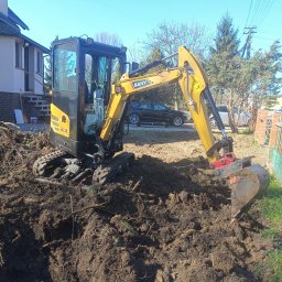 FPHU Damian Spychalski - Kompaktowa koparka SANY w trakcie prac ziemnych na posesji, widoczne wykopane bruzdy w ziemi, dom w tle. Słoneczny dzień, błękitne niebo.