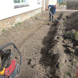 FPHU Damian Spychalski - Ręczne kopanie rowu przy budynku przez pracownika w kombinezonie. W tle widoczne ślady gąsienic koparki. Praca w toku, oświetlenie naturalne.