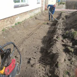 Ręczne kopanie rowu przy budynku przez pracownika w kombinezonie. W tle widoczne ślady gąsienic koparki. Praca w toku, oświetlenie naturalne.