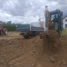 FPHU Damian Spychalski - Operator w minikoparce aktywnie kopie ziemię, tworząc świeży kopiec. W tle czerwony traktor, ciężarówka i nowoczesny dom pod pochmurnym niebem.