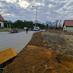 FPHU Damian Spychalski - Świeżo przygotowany teren z brązową ziemią obok brukowanej drogi, na której stoi pracownik. W tle biały bus, przyczepa z narzędziami, domy i zachmurzone niebo.