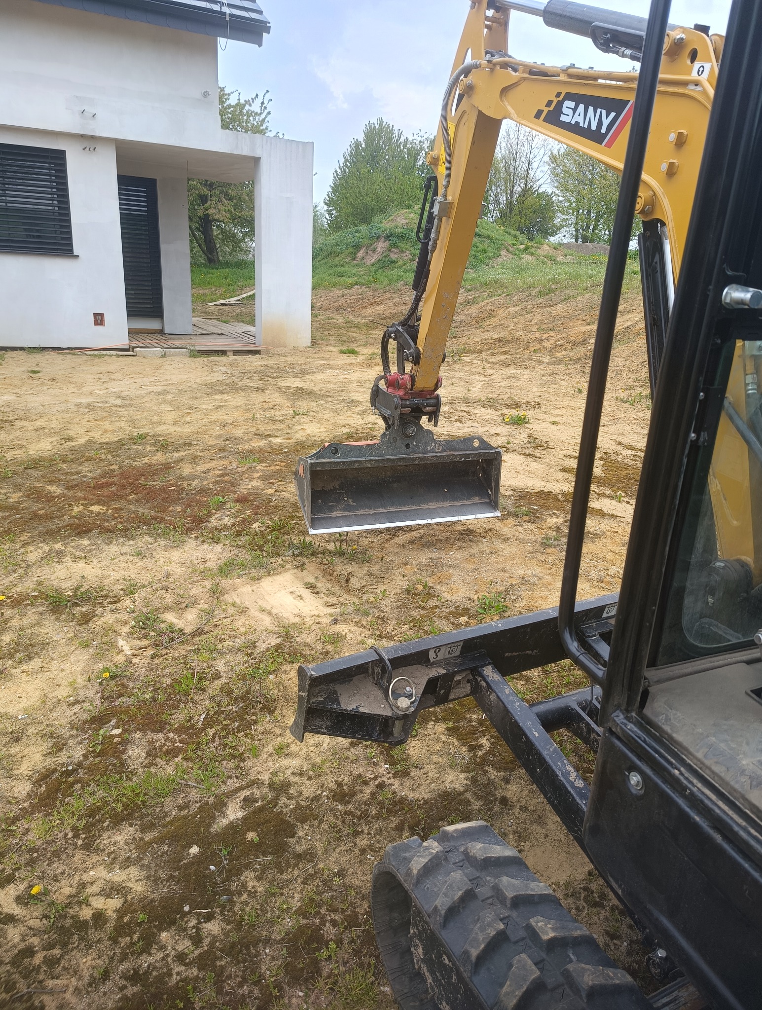 Żółta koparka SANY z łyżką na piaszczystym terenie budowy, widok z wnętrza kabiny. W tle biały, nowoczesny dom z czarnymi oknami i zielona roślinność.