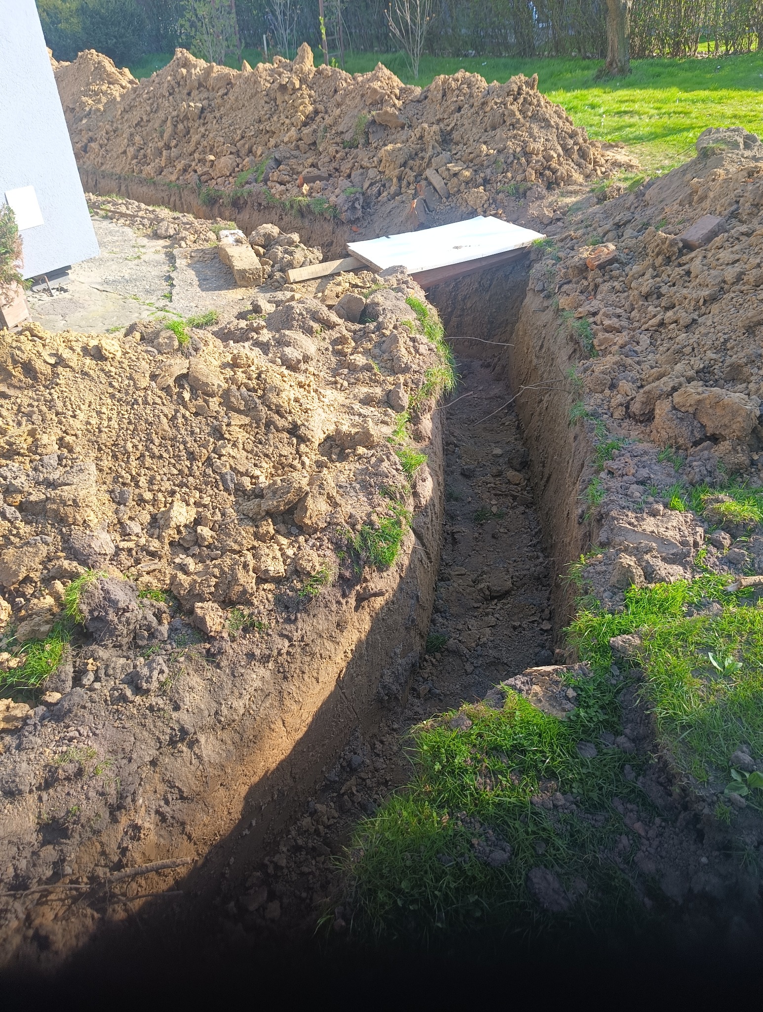 Wąski wykop w ziemi, zabezpieczony deskami, obok pryzma ziemi z zieloną trawą. Wykop pod instalacje w ogrodzie, w słoneczny dzień.