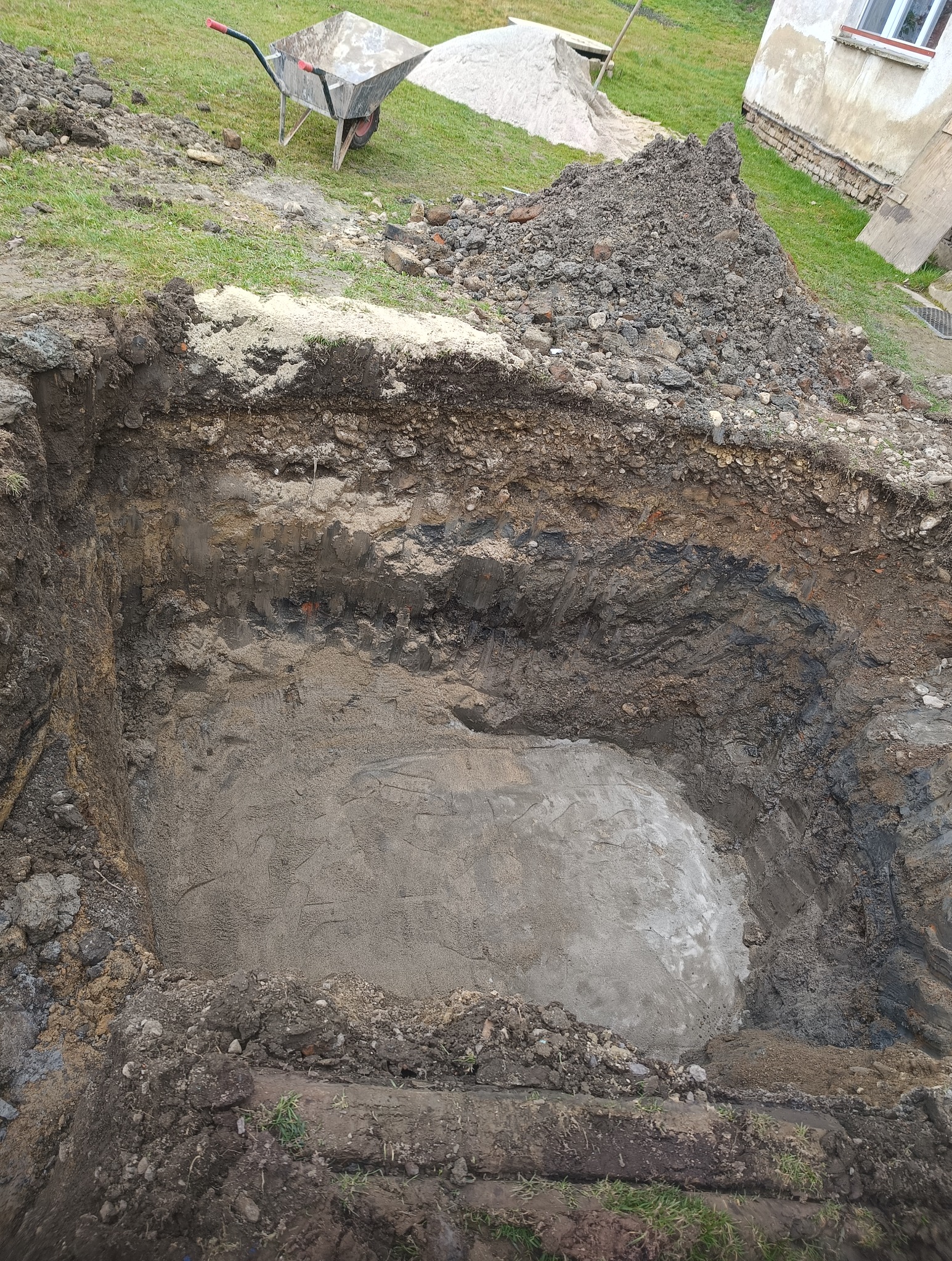 Wykopany dół w ziemi, widoczna warstwa piasku na dnie. Obok leży sterta ziemi i taczka. Etap prac ziemnych, prawdopodobnie pod fundamenty lub szambo.