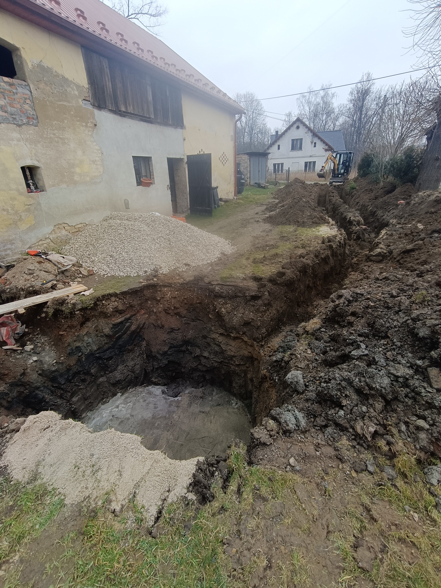 Głęboki wykop w ziemi obok starego budynku, z kupą żwiru i widocznym mini-koparką w tle. Etap prac ziemnych, przygotowanie pod fundamenty lub instalacje.