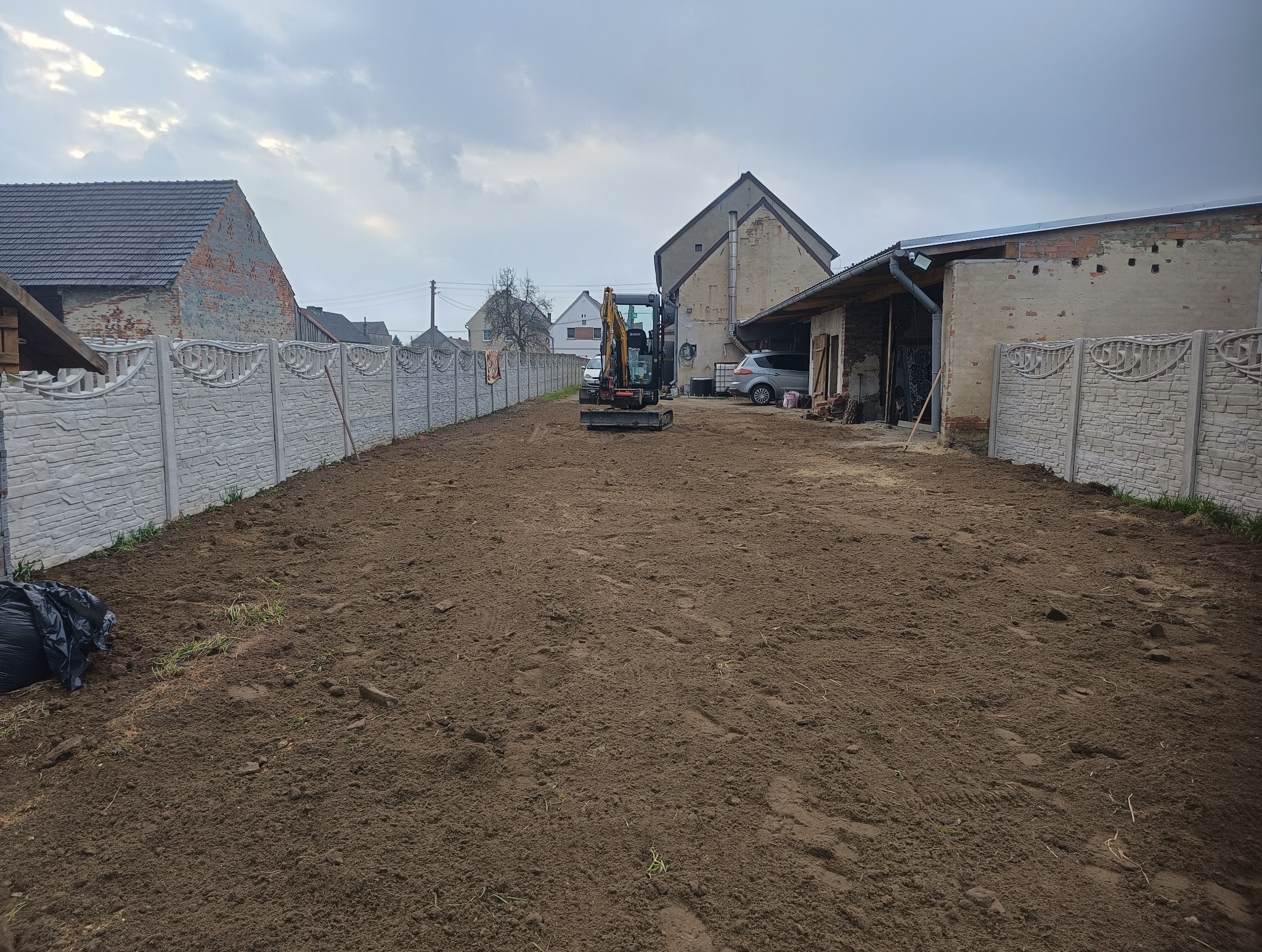 Mini koparka na działce z ziemią, w tle budynki i ogrodzenie z betonowych paneli. Przygotowanie terenu pod ogród. Widok na posesję i maszyny.