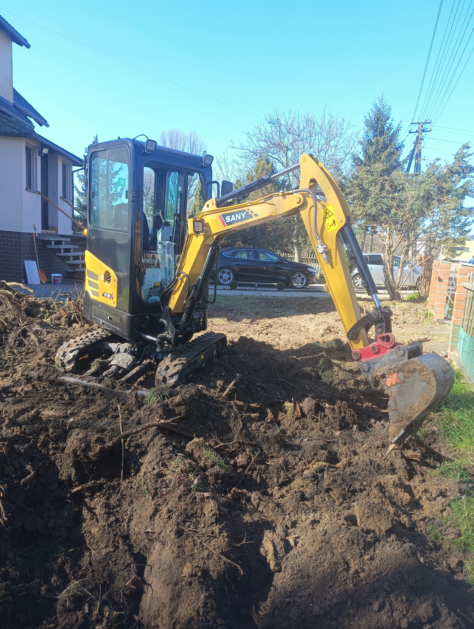 Kompaktowa koparka SANY w trakcie prac ziemnych na posesji, widoczne wykopane bruzdy w ziemi, dom w tle. Słoneczny dzień, błękitne niebo.