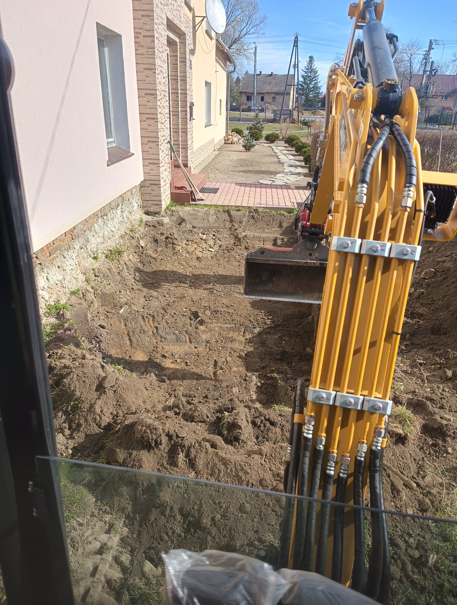 Żółta koparka wykonuje wykop wzdłuż fundamentów domu jednorodzinnego. Widok z kabiny operatora. Wykop przygotowany pod drenaż.