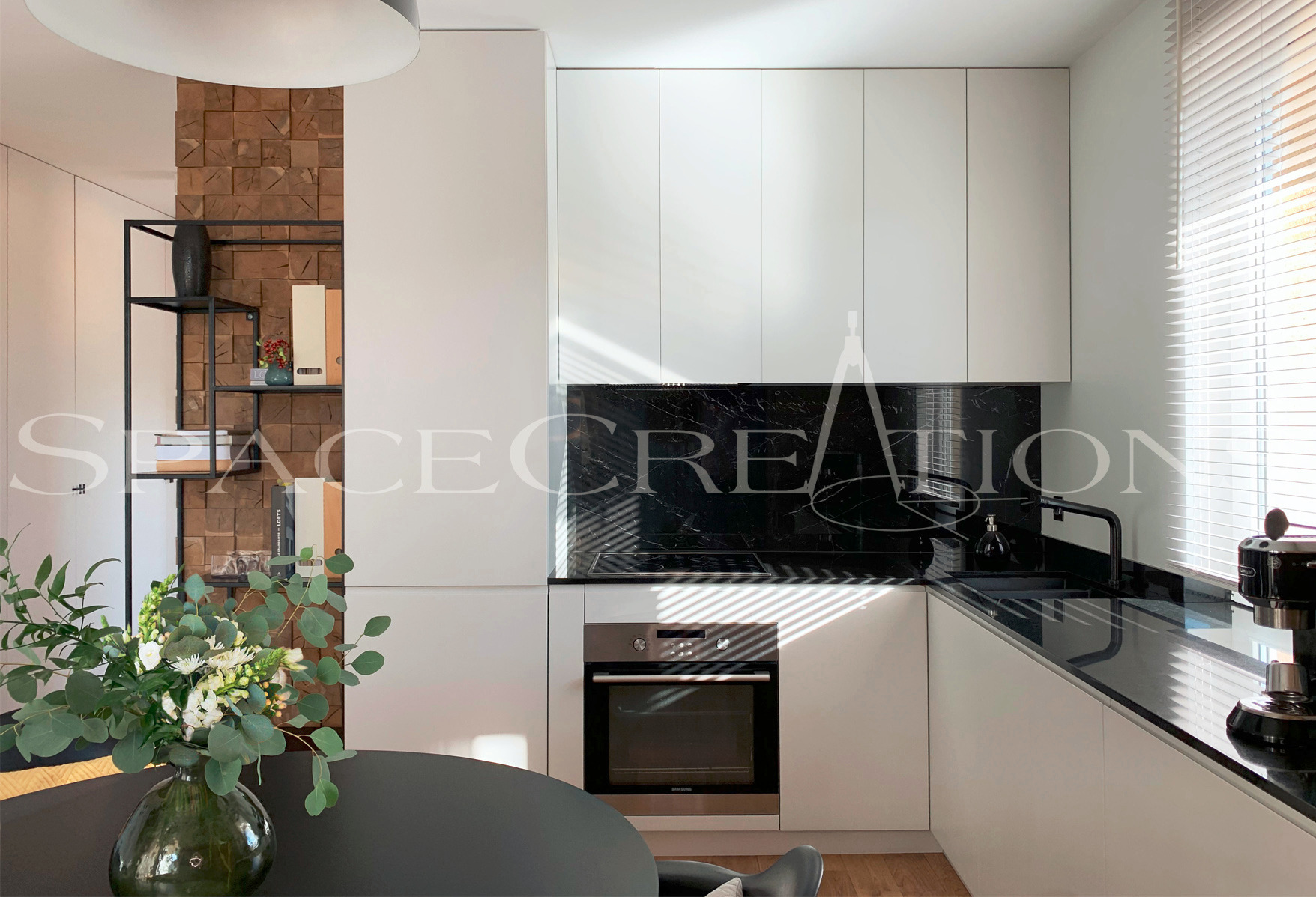 Nowoczesna kuchnia w bieli i czerni z drewnianym akcentem. Minimalistyczne szafki, czarny marmur na ścianie, okrągły stół z bukietem kwiatów. Oświetlenie sufitowe i żaluzje.
