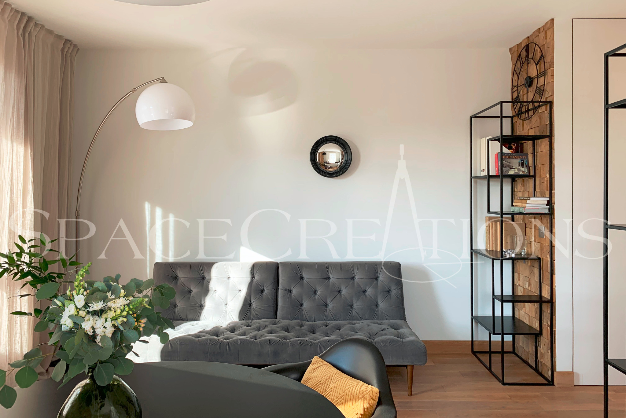 Stylowy salon z szarą sofą, okrągłym lustrem i metalowym regałem na tle ściany z drewnianą okładziną. Wazon z kwiatami na pierwszym planie, lampa łukowa.