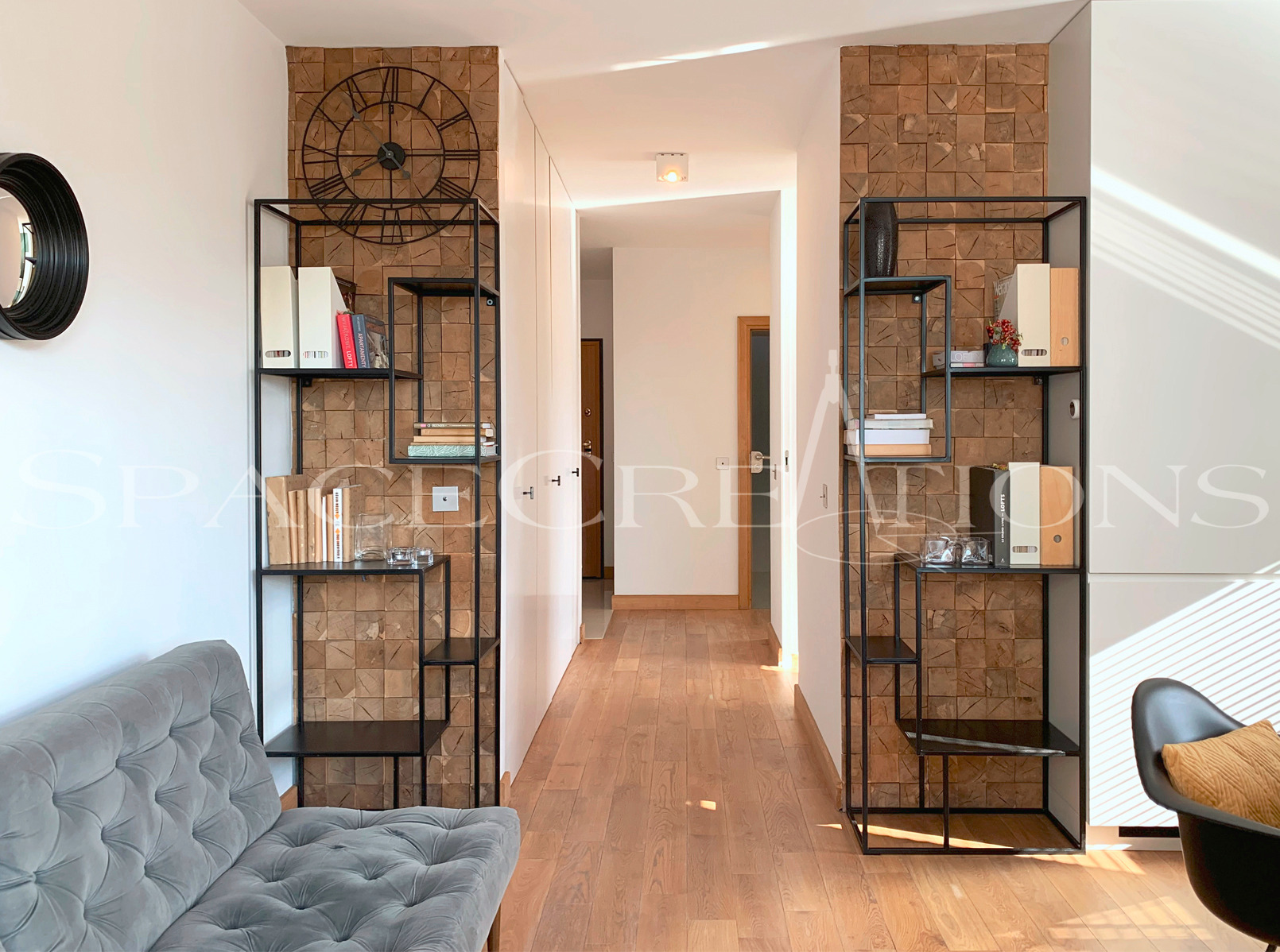 Wnętrze korytarza z drewnianą mozaiką na ścianie, metalowymi regałami i szarą sofą. Widok na jasne przejście z drewnianą podłogą i białymi ścianami.