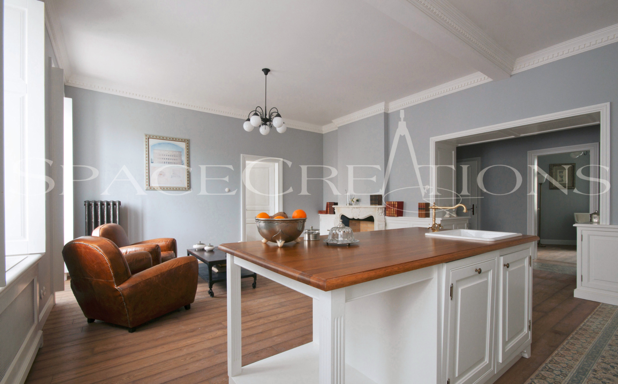 Jasne wnętrze z drewnianą podłogą i wyspą kuchenną z blatem z drewna. W tle widoczne skórzane fotele i dekoracyjny kominek. Całość w stylu klasycznym, z dbałością o detale.